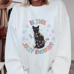May include: White sweatshirt with a black cat tangled in Christmas lights. The text "Is this jolly enough?" is printed in rainbow colors.
