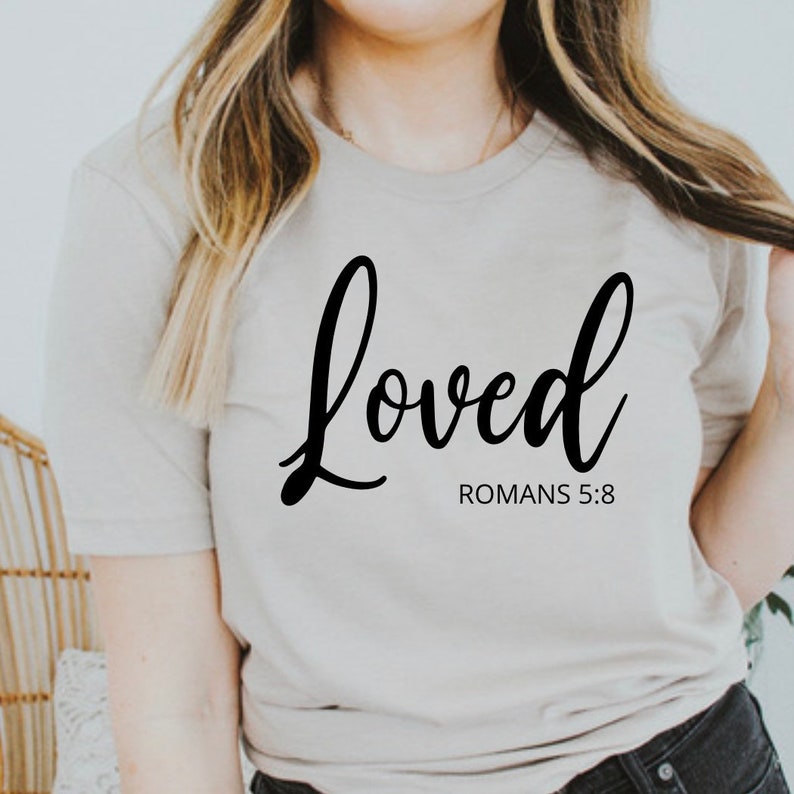 May include: A light gray t-shirt with the word "Loved" written in black cursive script. Below the word is the text "Romans 5:8" in black.