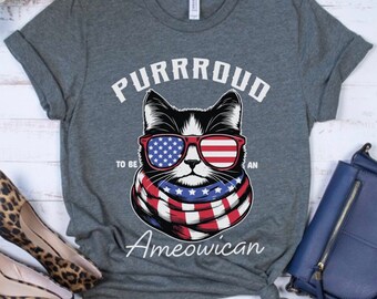 May include: A dark gray t-shirt with a black and white cat wearing red, white, and blue sunglasses and a scarf. The text "Purrroud to be an Ameowican" is printed on the shirt.