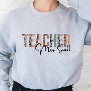 Pode incluir: Camisola cinzenta com a palavra "TEACHER" em estampado de leopardo e riscas coloridas. O texto "Mrs. Scott" est&aacute; escrito em it&aacute;lico abaixo da palavra "TEACHER".