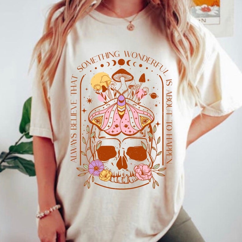 Op de afbeelding: Een cr&egrave;mekleurig T-shirt met een grafisch ontwerp met een schedel met een mot erboven, omringd door bloemen en paddenstoelen. De tekst "Always believe that something wonderful is about to happen" staat rondom het ontwerp.