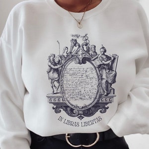 May include: White sweatshirt featuring a blue and white graphic print of a vintage illustration depicting a woman holding a scroll, a man with a crown, and a mythical creature. The text "IN LIBRAS LIBERTAS" is printed beneath the illustration.