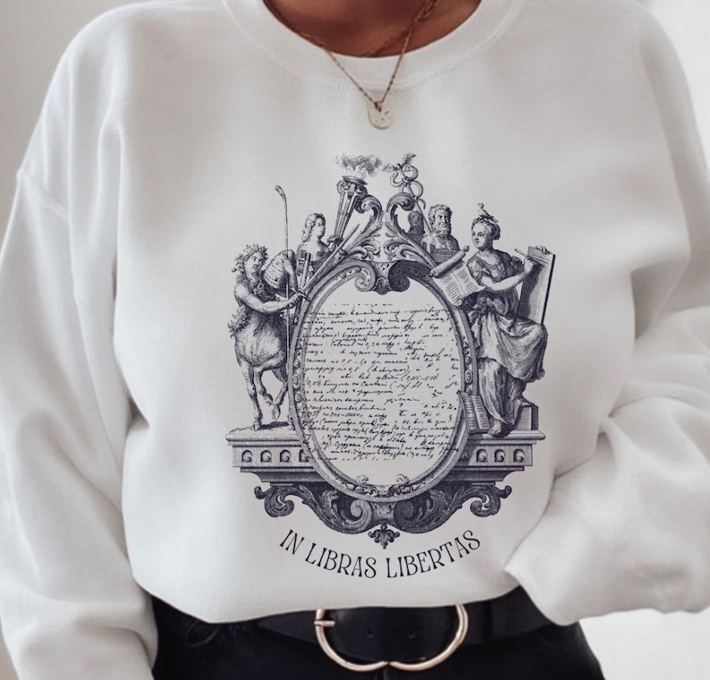May include: White sweatshirt featuring a blue and white graphic print of a vintage illustration depicting a woman holding a scroll, a man with a crown, and a mythical creature. The text "IN LIBRAS LIBERTAS" is printed beneath the illustration.