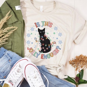 May include: A white t-shirt with a black cat wearing Christmas lights and the text "Is this jolly enough?" in colorful letters.