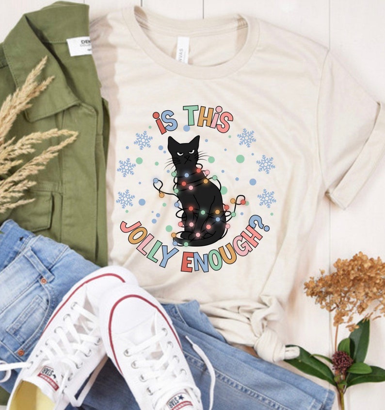 May include: A white t-shirt with a black cat wearing Christmas lights and the text "Is this jolly enough?" in colorful letters.