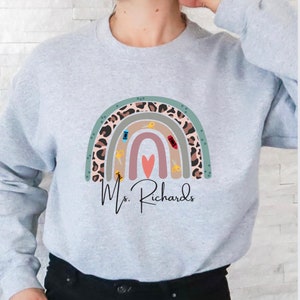 May include: A light gray sweatshirt featuring a colorful rainbow design with sewing-themed elements. The rainbow includes a leopard print pattern, scissors, and a heart. The name "Ms. Richards" is written below the rainbow in a script font.