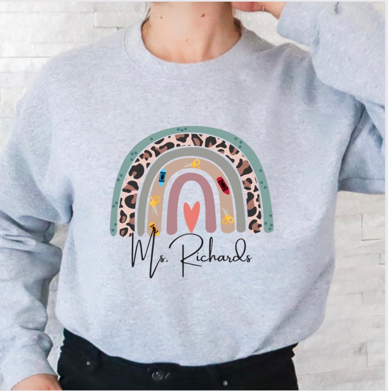May include: A light gray sweatshirt featuring a colorful rainbow design with sewing-themed elements. The rainbow includes a leopard print pattern, scissors, and a heart. The name "Ms. Richards" is written below the rainbow in a script font.