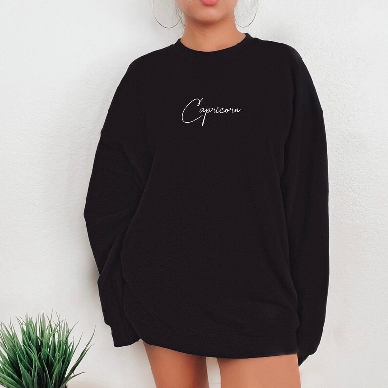 May include: Black sweatshirt with a white cursive inscription that reads "Capricorn".