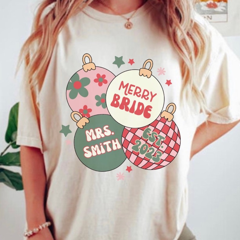 K&ouml;nnte beinhalten: Ein wei&szlig;es T-Shirt mit einem Weihnachtsbaumschmuck-Design. Die Ornamente sind rosa, gr&uuml;n und rot mit dem Text "Merry Bride", "Mrs. Smith" und "Est. 2023".