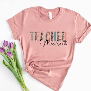 Pode incluir: Uma camiseta rosa com a palavra "TEACHER" em texto preto com um padr&atilde;o de leopardo. O texto "Mrs. Scott" est&aacute; escrito em it&aacute;lico preto abaixo da palavra "TEACHER".