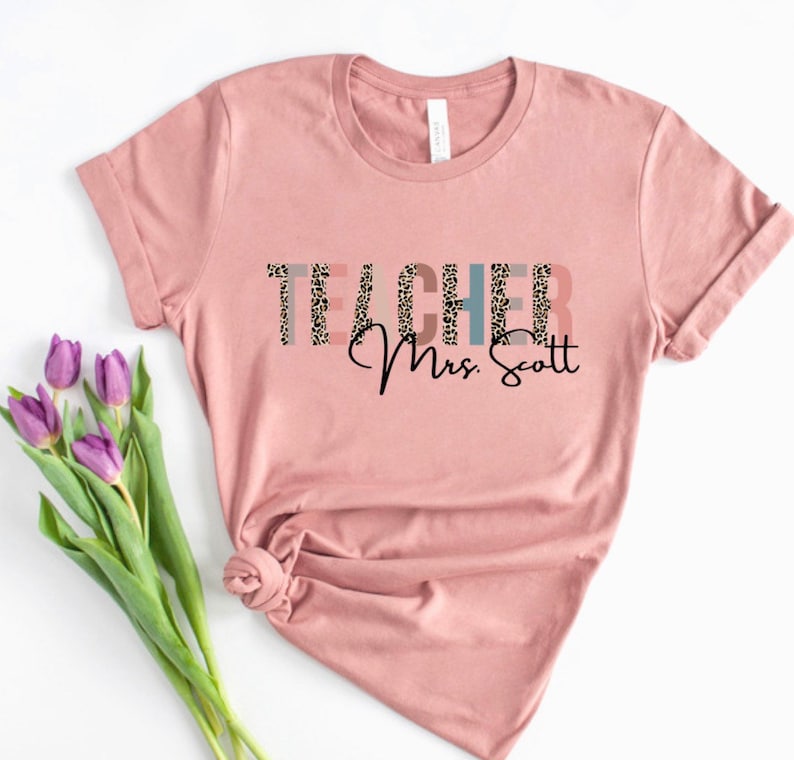 Pode incluir: Uma camiseta rosa com a palavra "TEACHER" em texto preto com um padr&atilde;o de leopardo. O texto "Mrs. Scott" est&aacute; escrito em it&aacute;lico preto abaixo da palavra "TEACHER".