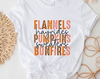 May include: White t-shirt with a graphic design that says "Flannels, Hayrides, Pumpkins, Sweaters, Bonfires" in brown and orange text.