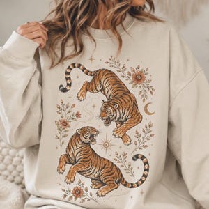 May include: A beige crewneck sweatshirt featuring a graphic print of two tigers in a dynamic pose. The tigers are surrounded by botanical illustrations of flowers, leaves, stars, and a crescent moon, all rendered in shades of orange, brown, and black.