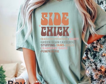 May include: A light blue t-shirt with the text "SIDE CHICK" in a retro font. The text is in shades of orange and brown. Below the text is a list of side dishes: mashed potatoes, gravy, green bean casserole, stuffing, yams, rolls, cranberry sauce, and pumpkin pie.