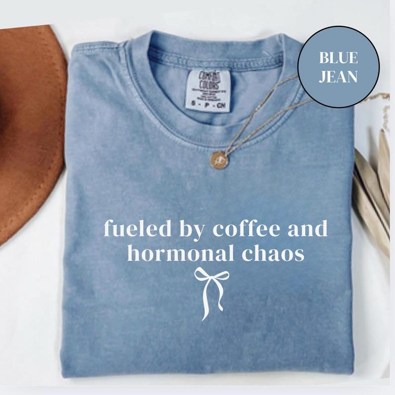 Puede incluir: Una camiseta azul vaquero con la frase "fueled by coffee and hormonal chaos" en escritura blanca. Un collar dorado con un colgante est&aacute; drapeado sobre la camisa. Un sombrero marr&oacute;n es parcialmente visible en la esquina superior izquierda.