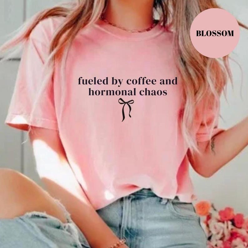 Puede incluir: Una camiseta rosa claro con el texto "fueled by coffee and hormonal chaos" y un lazo negro. La camiseta es de cuello redondo.