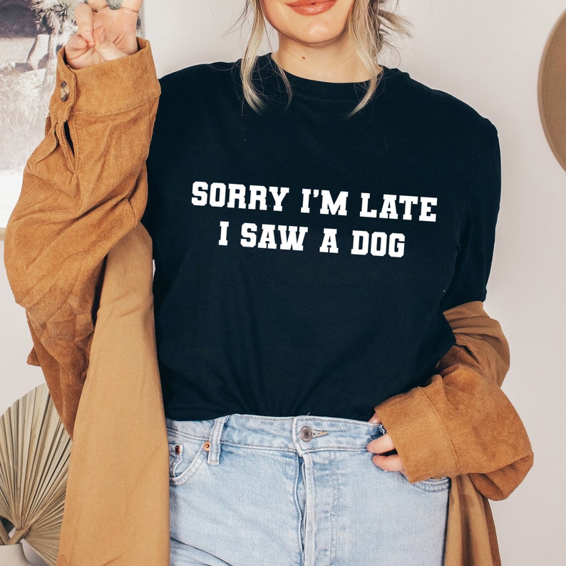 May include: A black t-shirt with white text that reads "Sorry I'm Late I Saw A Dog". The person wearing the shirt is also wearing blue jeans and a brown jacket.
