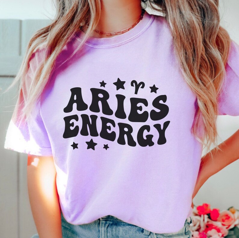 May include: A light purple sweatshirt with the words "ARIES ENERGY" in black, stylized font. The design includes stars and the Aries zodiac symbol. The sweatshirt is a soft, casual garment.