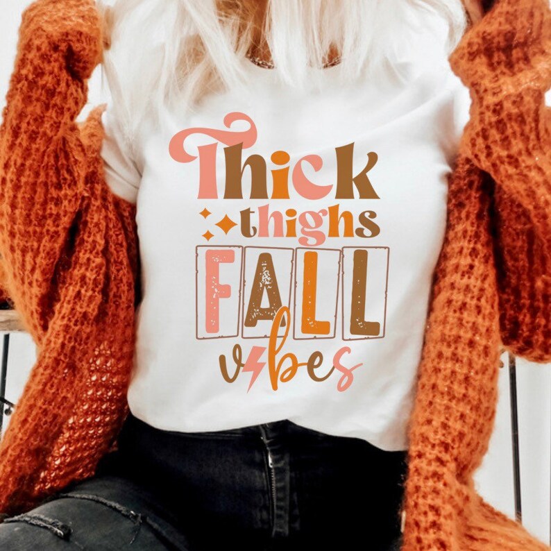 May include: White t-shirt with a graphic design that says "Thick thighs fall vibes" in brown, pink, and orange lettering.