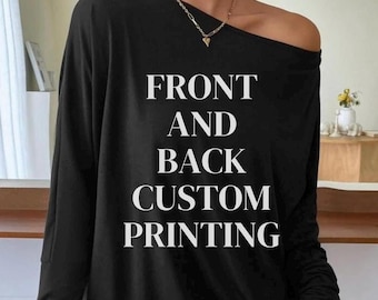 Op de afbeelding: Een zwart shirt met lange mouwen en een off-shoulder halslijn, voorzien van de witte tekst FRONT AND BACK CUSTOM PRINTING in het midden op de borst. Het shirt wordt gedragen met een dunne goudkleurige ketting met een klein hartvormig hangertje.