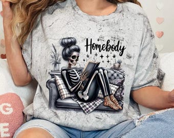 May include: A gray t-shirt with a skeleton wearing a messy bun, reading a book on a couch. The text "Homebody" is printed on the shirt.