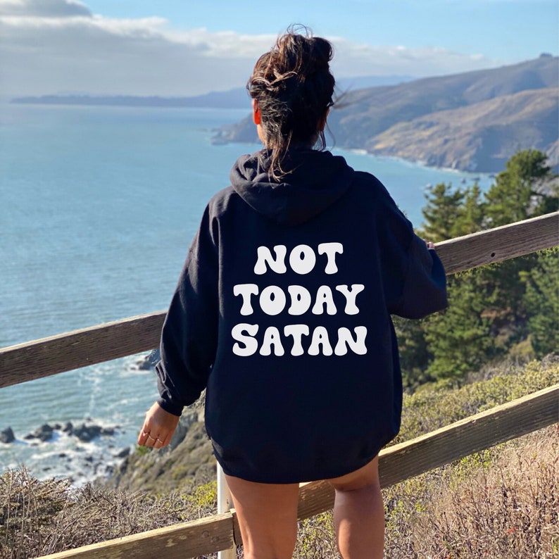 K&ouml;nnte beinhalten: Ein schwarzer Hoodie mit einem wei&szlig;en Aufdruck, der in einer Retro-Schrift "NOT TODAY SATAN" sagt. Der Hoodie wird von einer Person getragen, die auf einem Holzgel&auml;nder steht und auf eine Wasserfl&auml;che blickt.