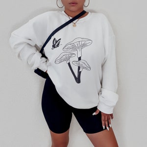 May include: White sweatshirt with a black mushroom graphic and a butterfly design. The sweatshirt has long sleeves and a crew neck.