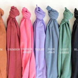 May include: A collection of seven folded t-shirts in various colors, including yam, crimson, crunch berry, violet, blue jean, light green, and black. Each shirt has a label with its color name.