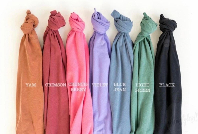 May include: A collection of seven folded t-shirts in various colors, including yam, crimson, crunch berry, violet, blue jean, light green, and black. Each shirt has a label with its color name.
