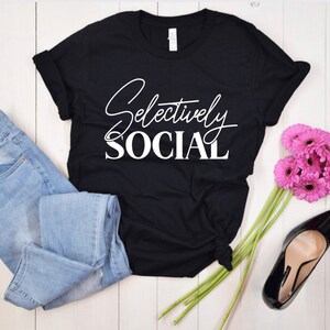 May include: Black t-shirt with white text that reads "Selectively Social". The shirt is folded and laid on a white wooden surface with a pair of blue jeans, a bouquet of pink flowers, and a black high heel shoe.