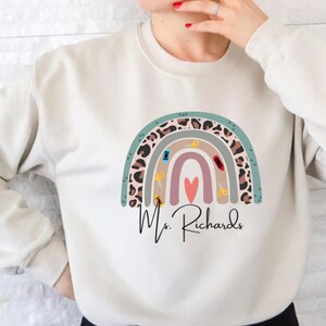 May include: Cream-colored sweatshirt featuring a colorful rainbow design with a leopard print accent. The rainbow includes scissors, a pencil, and a heart. The name "Ms. Richards" is written below the rainbow in a script font.