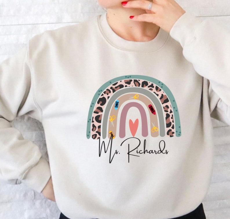May include: Cream-colored sweatshirt featuring a colorful rainbow design with a leopard print accent. The rainbow includes scissors, a pencil, and a heart. The name "Ms. Richards" is written below the rainbow in a script font.