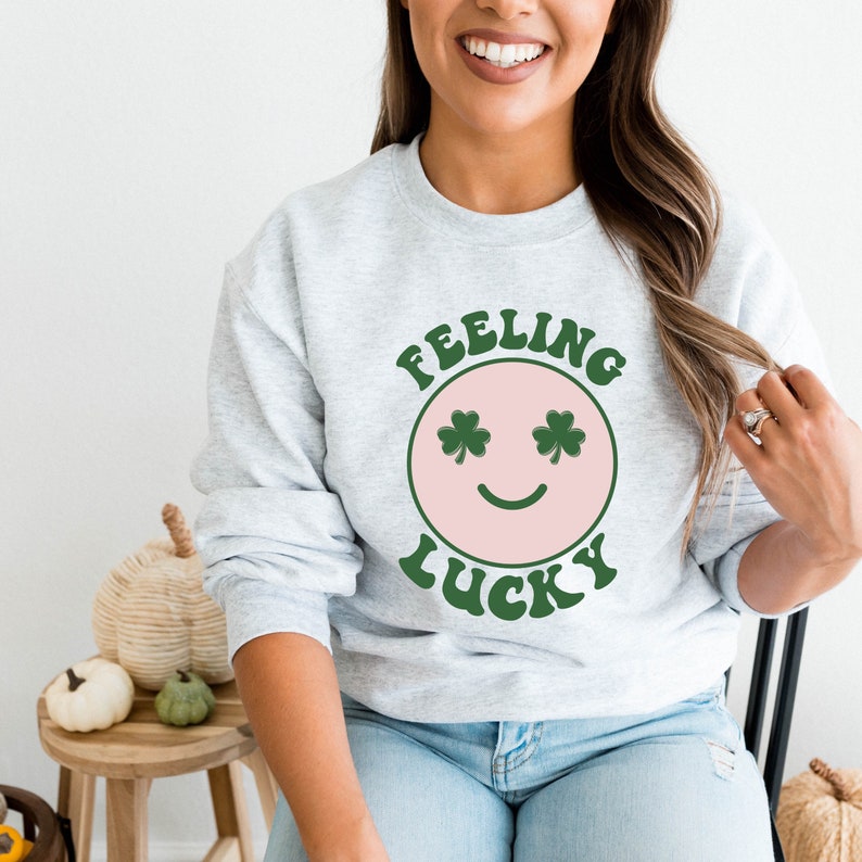 Puede incluir: Una sudadera gris con una cara sonriente rosa con ojos de tr&eacute;bol verde. El texto "Feeling Lucky" est&aacute; escrito en verde arriba y abajo de la cara sonriente.