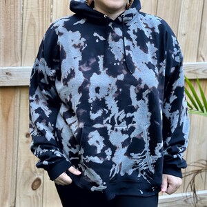May include: A black hooded sweatshirt featuring a bleached, mottled pattern in shades of grey and charcoal. The garment includes a front kangaroo pocket, long sleeves with ribbed cuffs, and a drawstring hood with black cords for an adjustable fit.