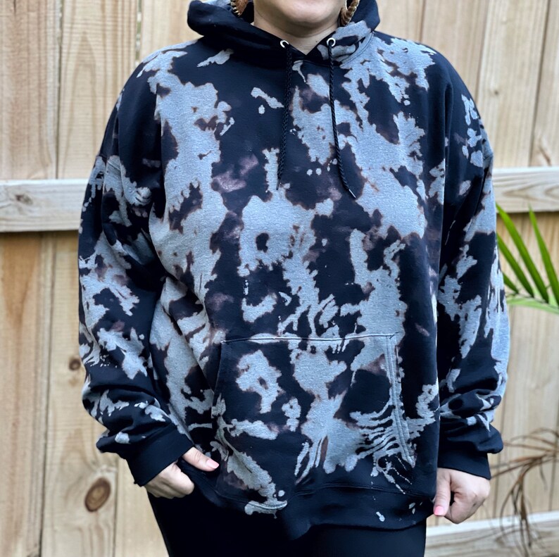 May include: A black hooded sweatshirt featuring a bleached, mottled pattern in shades of grey and charcoal. The garment includes a front kangaroo pocket, long sleeves with ribbed cuffs, and a drawstring hood with black cords for an adjustable fit.