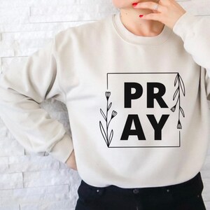 May include: A white sweatshirt with the word "PRAY" in black lettering inside a square frame. The frame is decorated with black floral designs.