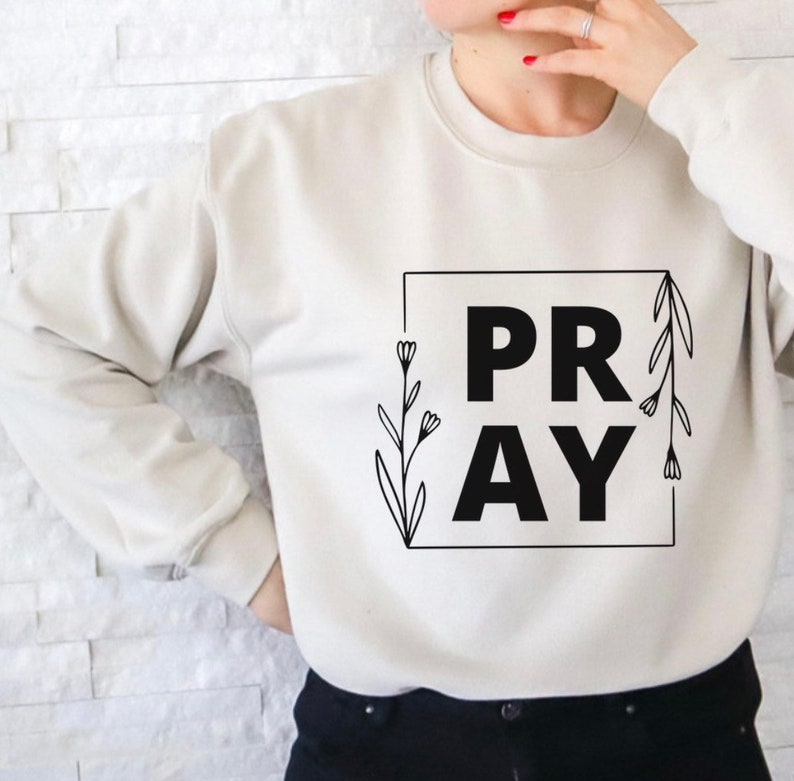 May include: A white sweatshirt with the word "PRAY" in black lettering inside a square frame. The frame is decorated with black floral designs.