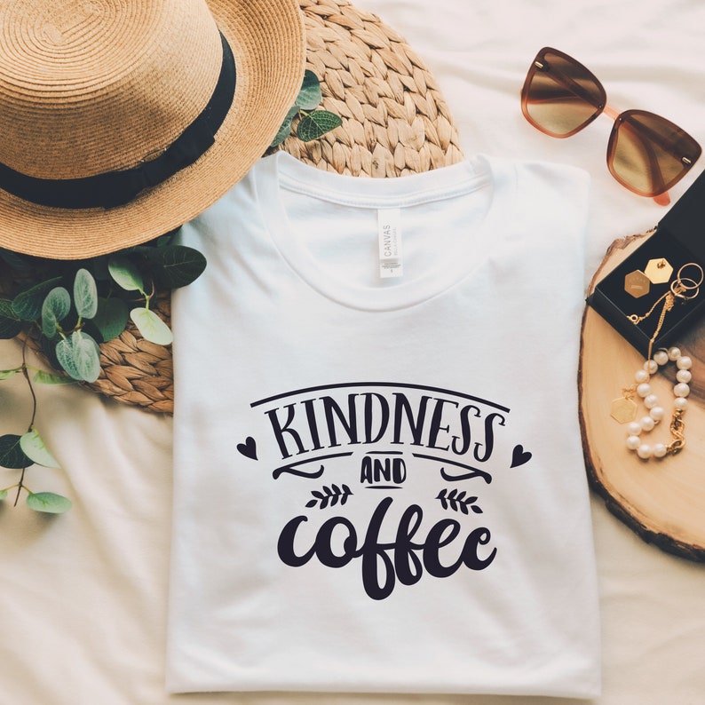 May include: White t-shirt with the text "Kindness and coffee" in black lettering with a heart on each side of the text and a small leaf design below the text.