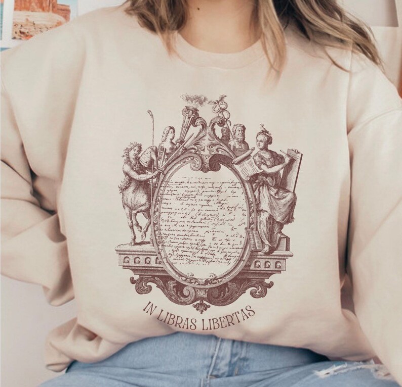 May include: A beige sweatshirt with a brown graphic print of a vintage illustration featuring a crest with figures and the text "IN LIBRAS LIBERTAS".