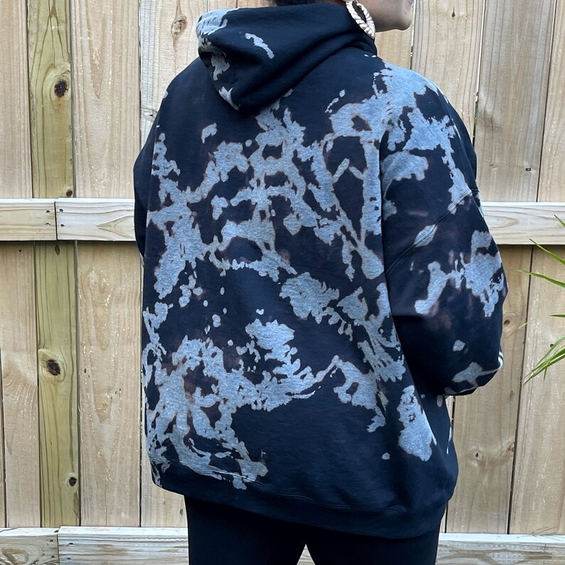 May include: A black hooded sweatshirt featuring a bleached, mottled pattern in shades of light gray and faded brown. The long-sleeved pullover garment includes a hood and a ribbed hem, displaying an irregular, abstract design across the back.