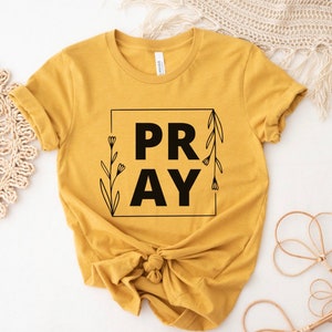 May include: A mustard yellow t-shirt with a black square graphic that says "PRAY" with floral accents.