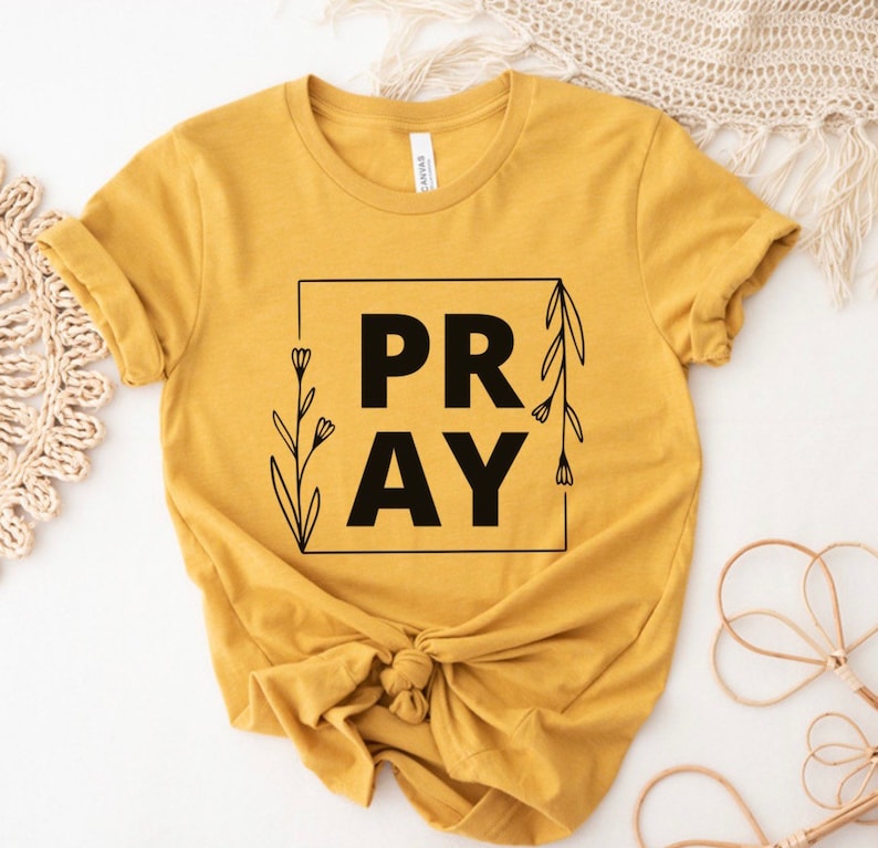 May include: A mustard yellow t-shirt with a black square graphic that says "PRAY" with floral accents.