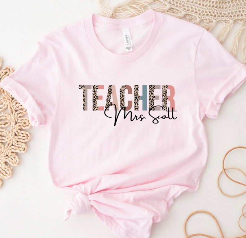 Pode incluir: Camiseta rosa com a palavra "TEACHER" em letras coloridas com um padr&atilde;o de leopardo. O texto "Mrs. Scott" est&aacute; escrito em it&aacute;lico abaixo da palavra "TEACHER".