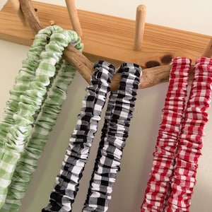 May include: Close-up of three fabric scrunchies in green, black and white, and red gingham patterns. The scrunchies are hanging on a wooden rack with natural wood pegs and a branch. The scrunchies are made of a soft, gathered fabric.