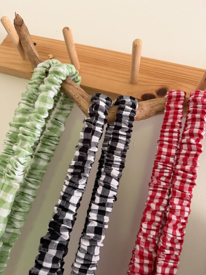 May include: Close-up of three fabric scrunchies in green, black and white, and red gingham patterns. The scrunchies are hanging on a wooden rack with natural wood pegs and a branch. The scrunchies are made of a soft, gathered fabric.
