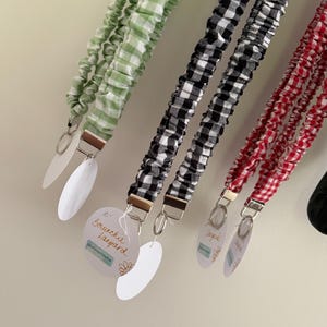 May include: A collection of scrunchie lanyards in green, black, and red gingham patterns. Each lanyard has a metal clasp and a white oval tag. The text "Scrunchie Lanyard" is visible on one tag.