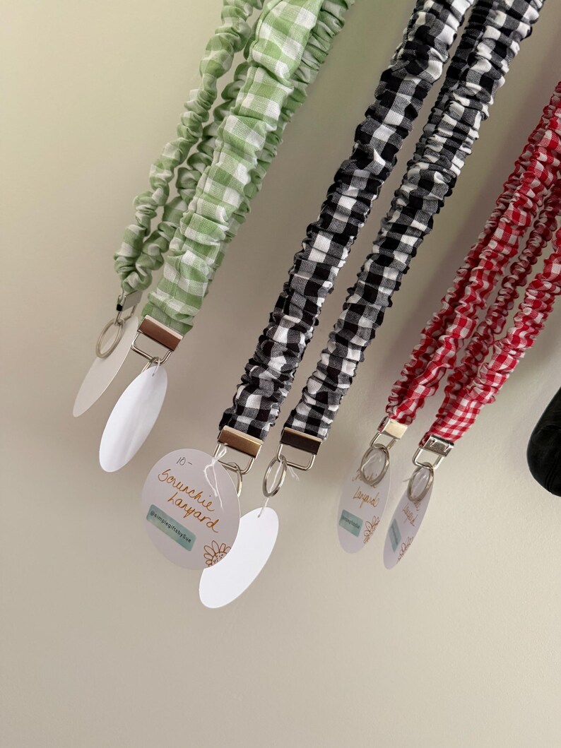 May include: A collection of scrunchie lanyards in green, black, and red gingham patterns. Each lanyard has a metal clasp and a white oval tag. The text "Scrunchie Lanyard" is visible on one tag.