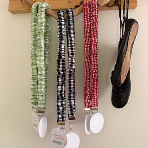 May include: Three scrunchie lanyards in green and white, black and white, and red and white gingham patterns hang from a wooden rack. Each lanyard has a metal clip and a white tag. A black shoe hangs on the right.