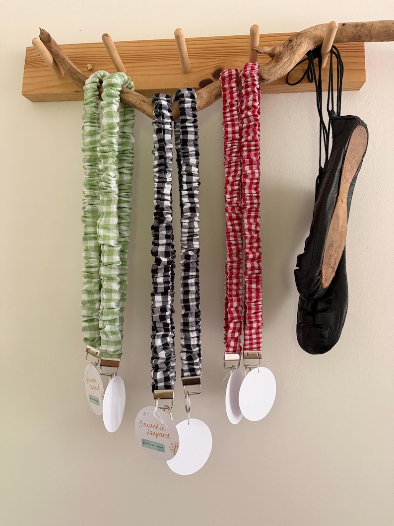 May include: Three scrunchie lanyards in green and white, black and white, and red and white gingham patterns hang from a wooden rack. Each lanyard has a metal clip and a white tag. A black shoe hangs on the right.