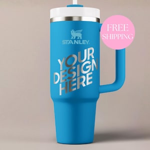 May include: A bright blue insulated tumbler with a handle, a white lid, and a straw. The Stanley logo is printed in white near the top. The text "YOUR DESIGN HERE" is printed in large silver letters across the center of the blue body.